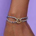 Multicolored braided bracelet with a gold ring on a wrist against a purple background