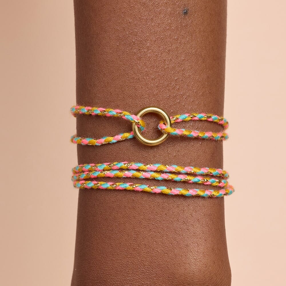 Multicolored braided bracelet with a gold ring on a wrist against a beige background